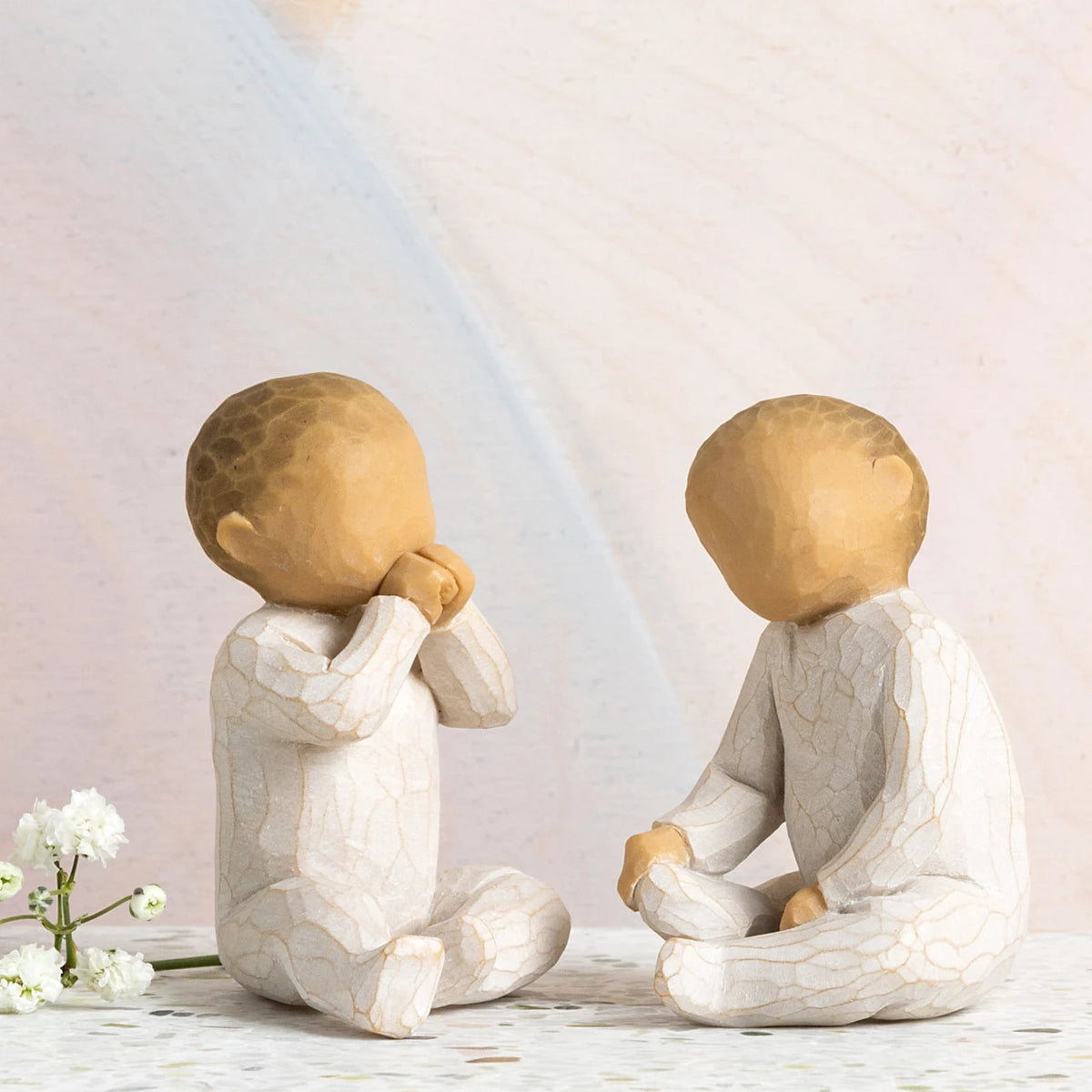 Willow Tree Two Together, two hand-painted seated resin figures of babies, tallest 2", one with hands at mouth, one with hands on legs, both in cream onsies, in front of pastel painting