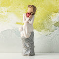 Willow Tree Always, 6”h hand-painted figure in cream dress, knees drawn up to chest, holding red heart to face, seated on gray rock etched with symbols of love, in front of lime painting