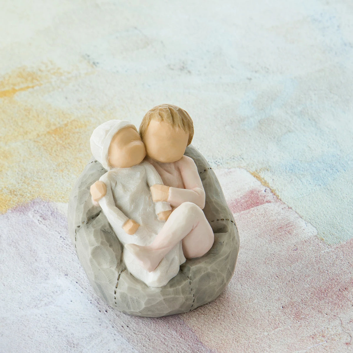 Willow Tree My new baby (blush), 2.5”h hand-painted resin figure of toddler in blush pink onesie holding infant in cream, cuddled together in a beanbag, in front of pastel painting