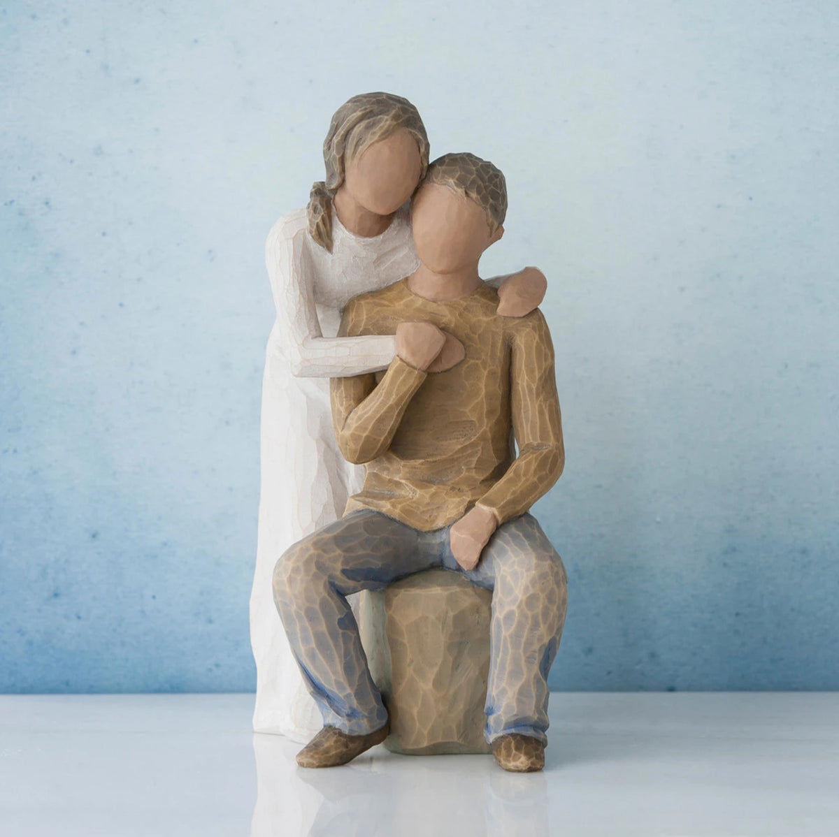 Man figurine sitting on rock wearing brown shirt and blue jeans with woman figurine in white dress with arms wrapped around him - blue background