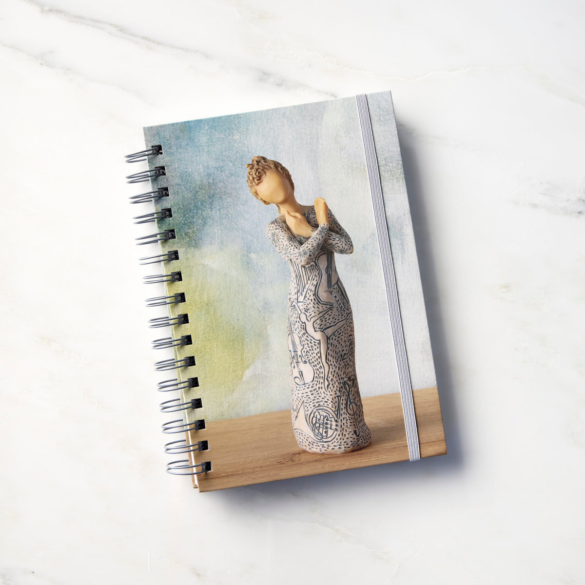 Front view: Spiral-bound notebook featuring front of female figure carved with contrasting pattern of dark blue musical symbols, instruments, dancing forms