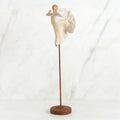 Far shot of angel figurine dressed in white dress dancing with arms and legs out - she is on a brown wooden stand