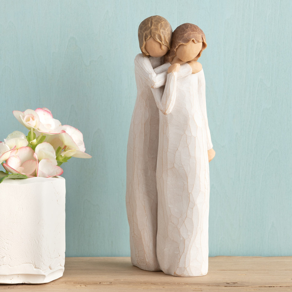Willow Tree Chrysalis, 9”h hand-painted standing resin figure of two women in cream dresses; one standing behind and embracing the other, on wooden shelf with flowers