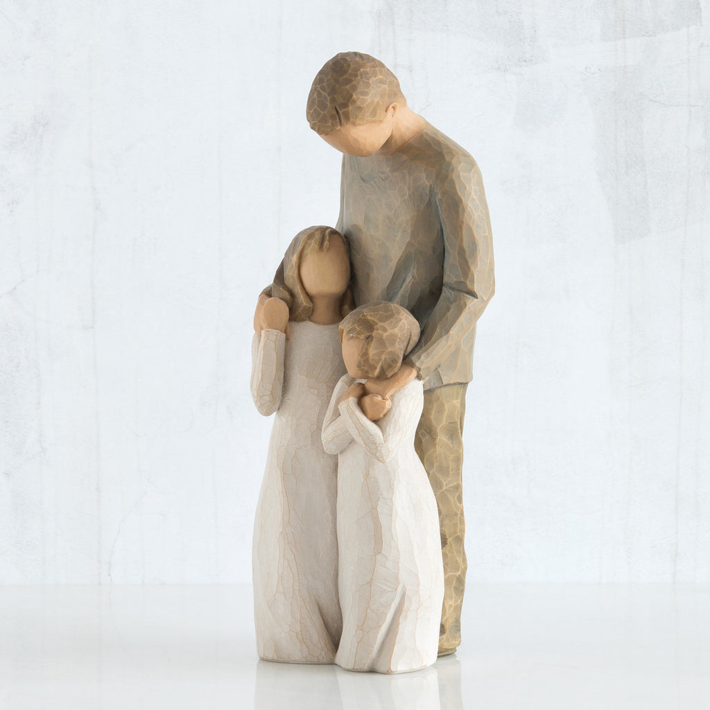 Man figurine in brown standing with arms around two little girl figurines in white