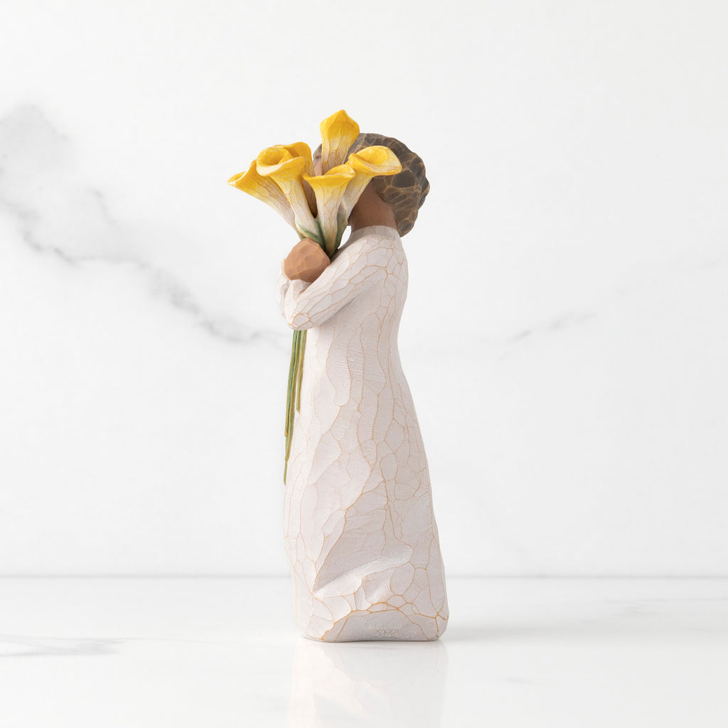 Left side of standing figure in cream dress with darker skin holding yellow calla lilies in her arms