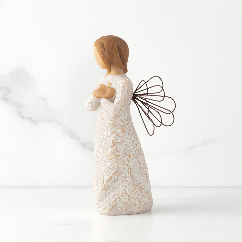 Left side view of standing female figure with wire wings in cream dress carved with natural symbols, hands crossed under gold heart on chest