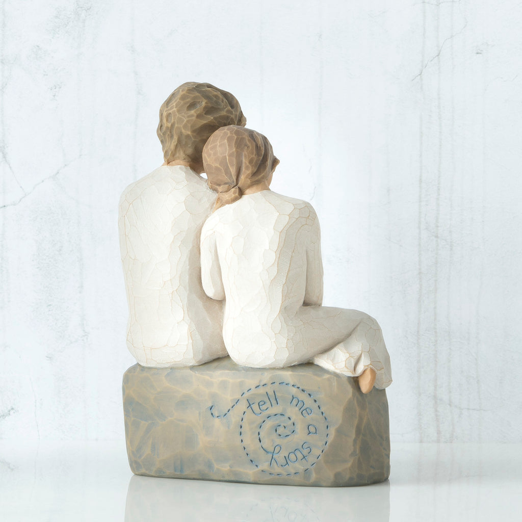 Back view of boy and girl figurine in white sitting on grey rock that says 'tell me a story' in blue carved in