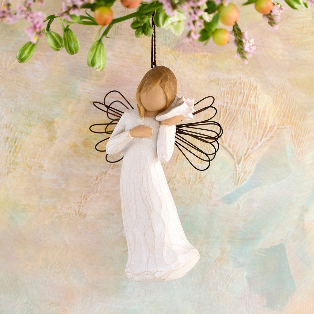 Willow Tree Thinking of You Ornament, 4.5”h hand-painted resin angel figure in cream dress with wire wings, holding pink conch shell to ear, brown elastic cord, hanging in front of peach painting