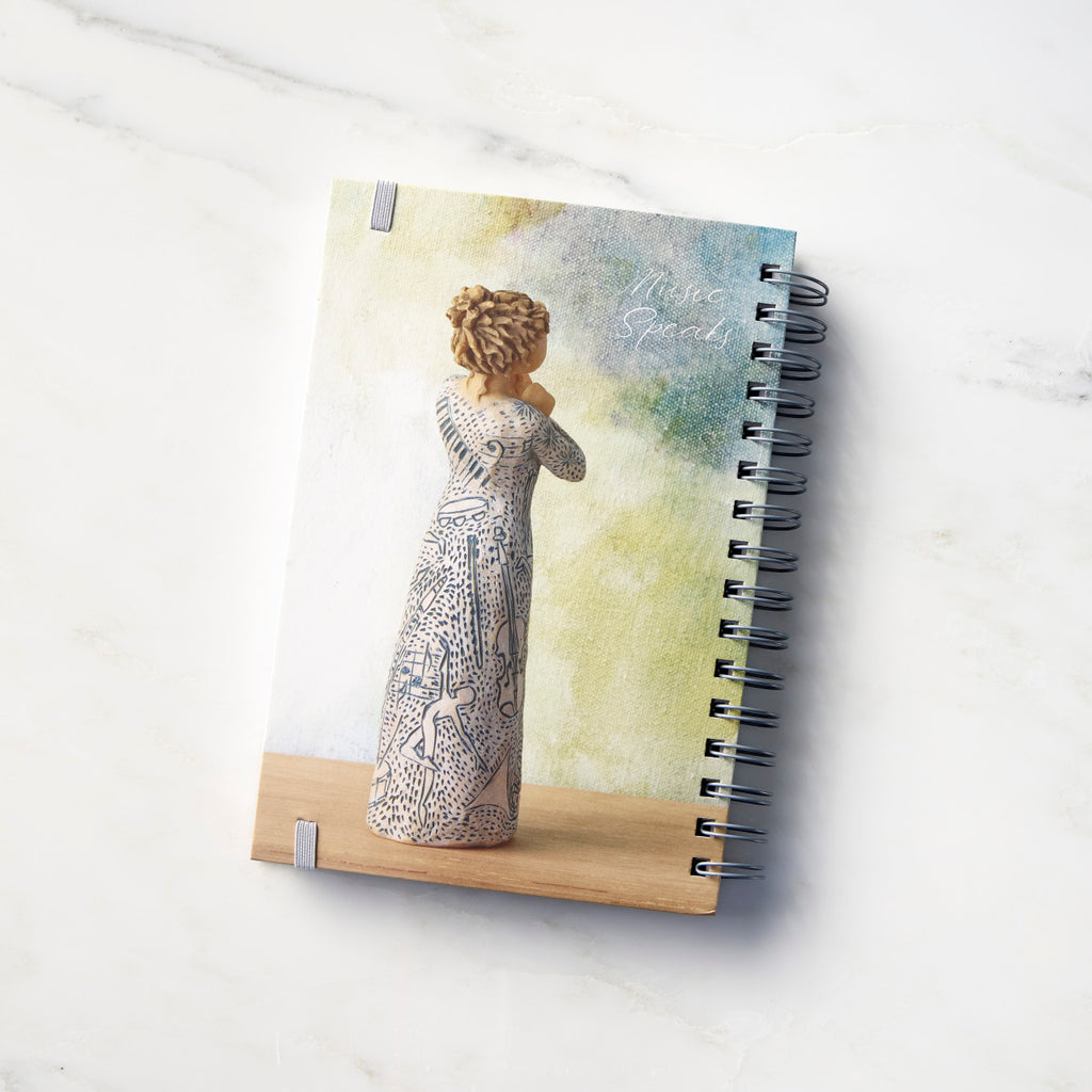 Back view: Spiral-bound notebook featuring back of female figure carved with contrasting pattern of dark blue musical symbols, instruments, dancing forms