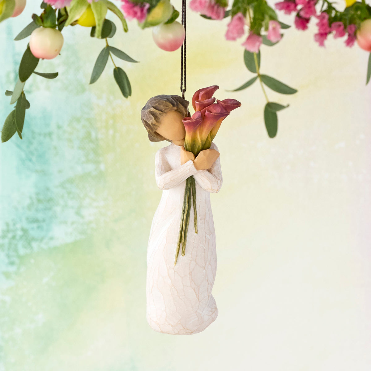 Willow Tree Bloom Ornament, 4.5”h hand-painted resin figure in cream dress, holding bouquet of fuchsia calla lilies, brown elastic cord, hanging in front of pale green painting