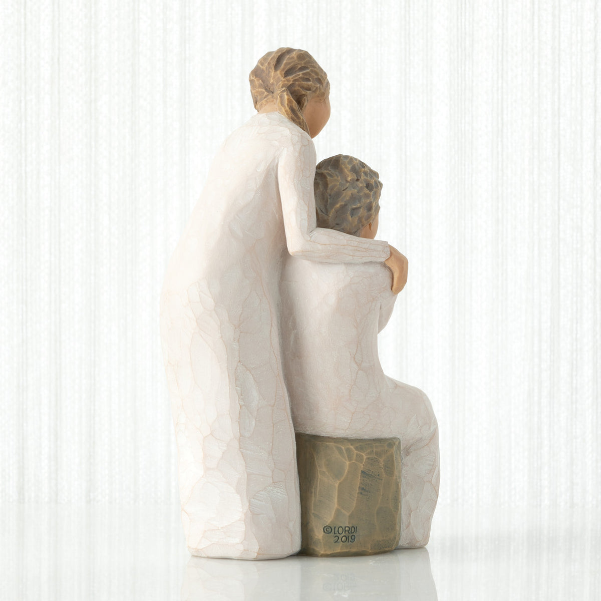 Back view, seated older female in cream dress, with younger female in cream dress, standing at her shoulder, hands embracing