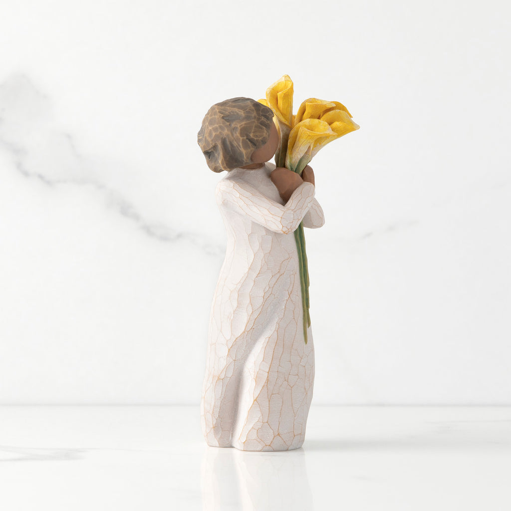 Right side of standing figure in cream dress with darker skin holding yellow calla lilies in her arms