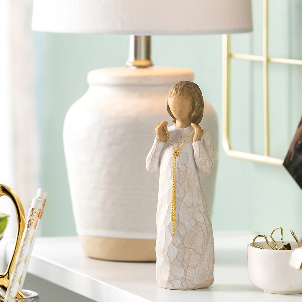 Willow Tree Truly Golden, 5"h hand-painted standing resin figure in cream dress, holding a knotted, long gold chain between her hands, on table with cream lamp