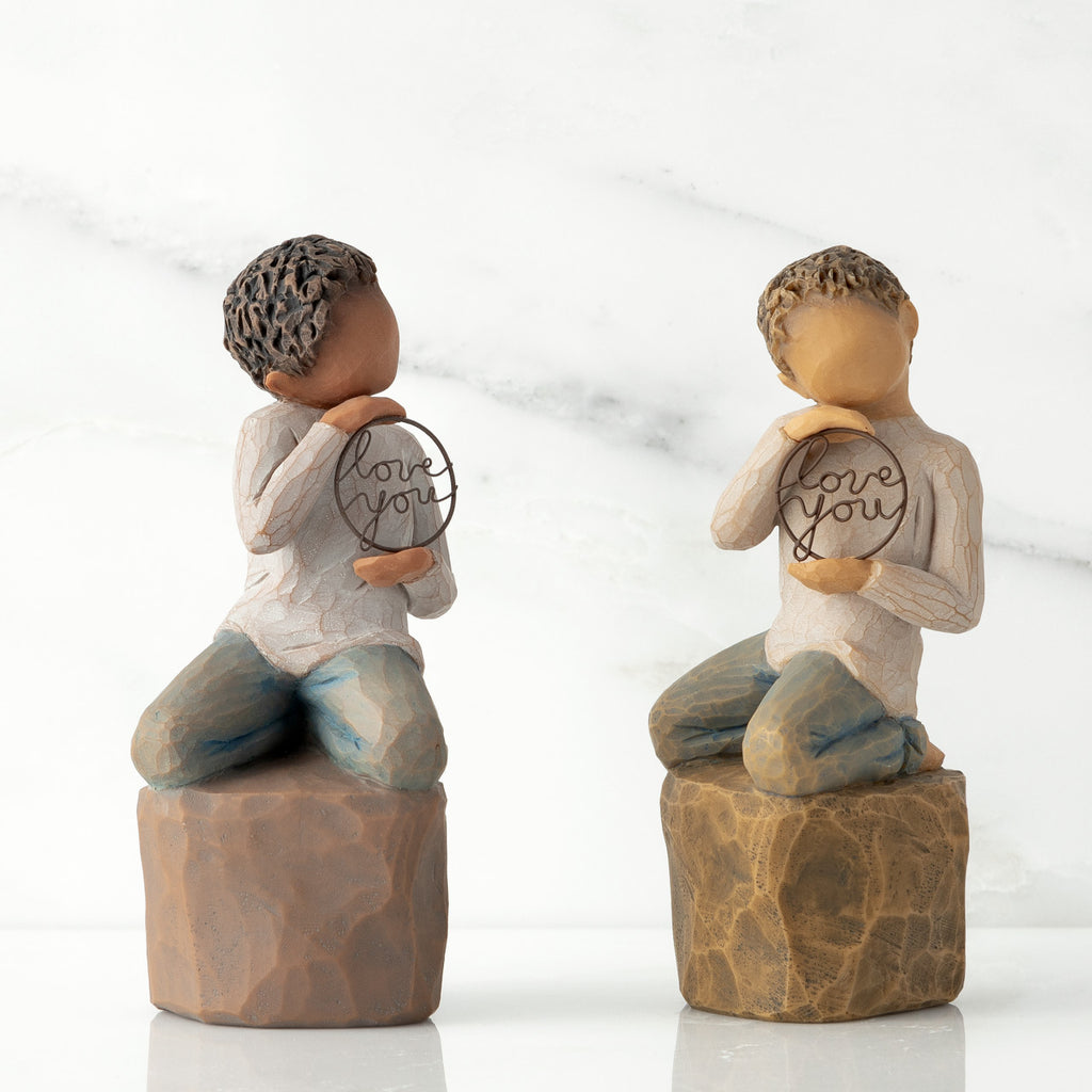 Two options (darker skin and lighter skin) of boy figurine kneeling on rock, holding wire circle with ‘love you’ inside