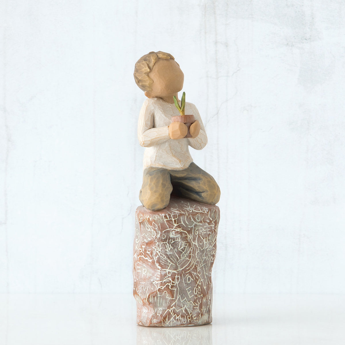 Front view of boy with brown hair sitting on a stump while holding a small potted plant to his chest