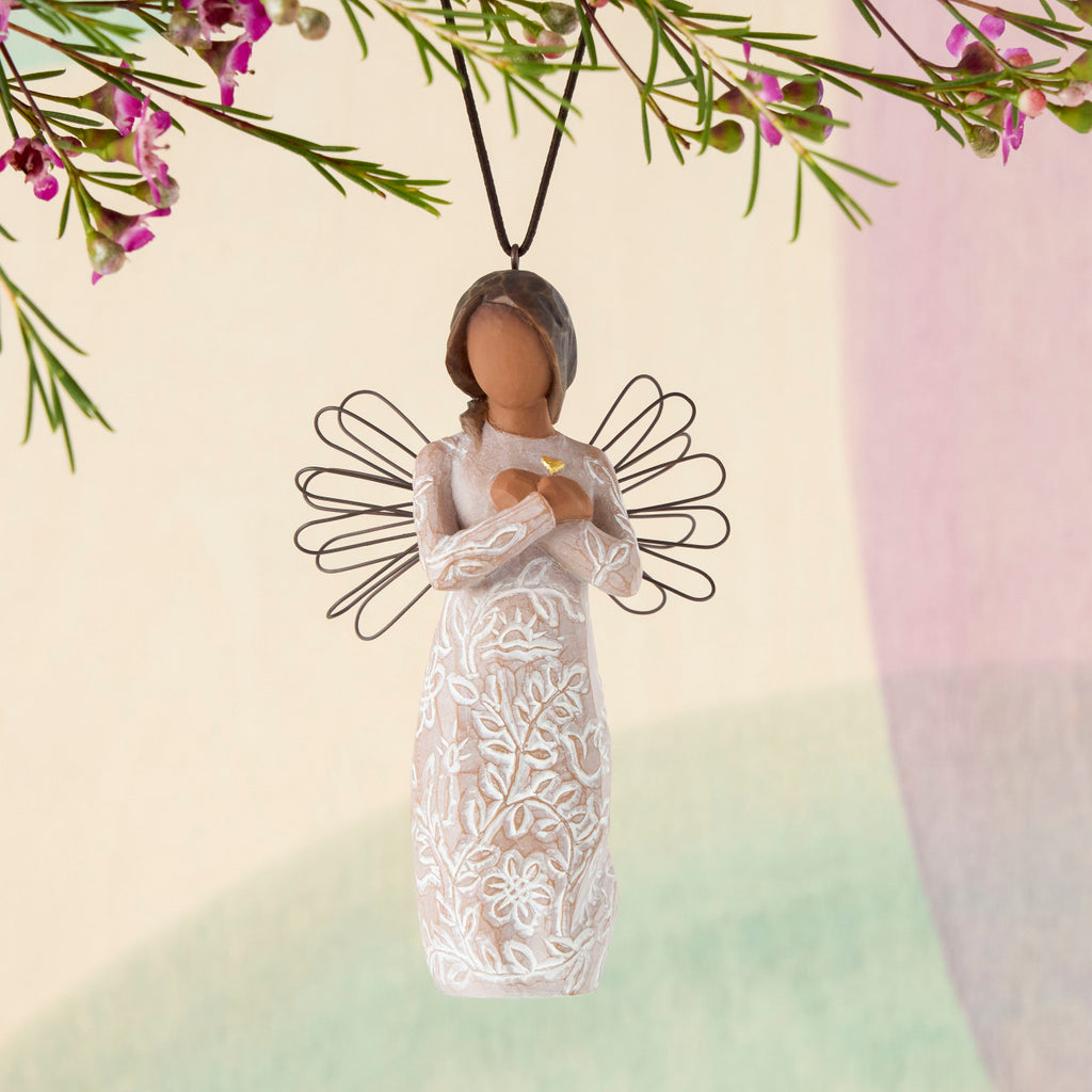 Willow Tree Remembrance Ornament, 4”h hand-painted resin angel figure with darker skin and hair, wire wings, arms crossed over gold heart, cream dress carved with nature imagery, brown elastic cord, hanging in front of pastel painting