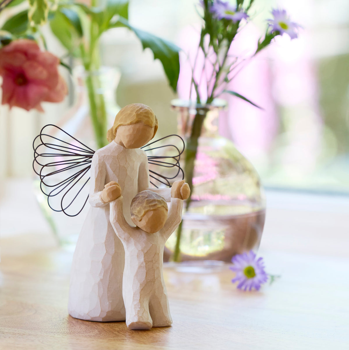Willow Tree Guardian Angel, 5”h hand-painted resin figure in cream dress and wire wings, holding hands of toddler in cream onesie walking ahead, on table with wildflowers
