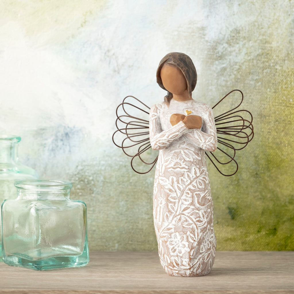 Willow Tree Remembrance (darker skin), 5.5”h hand-painted standing resin angel figure with wire wings, arms crossed over gold heart, in cream dress carved with nature icons, in front of grey-green painting