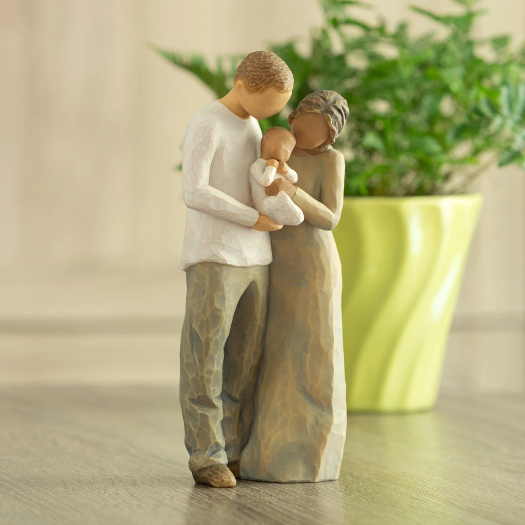 Front view of standing couple, woman has darker skin and hair; man has lighter skin and hair, holding a baby between them. Potted plant in background.