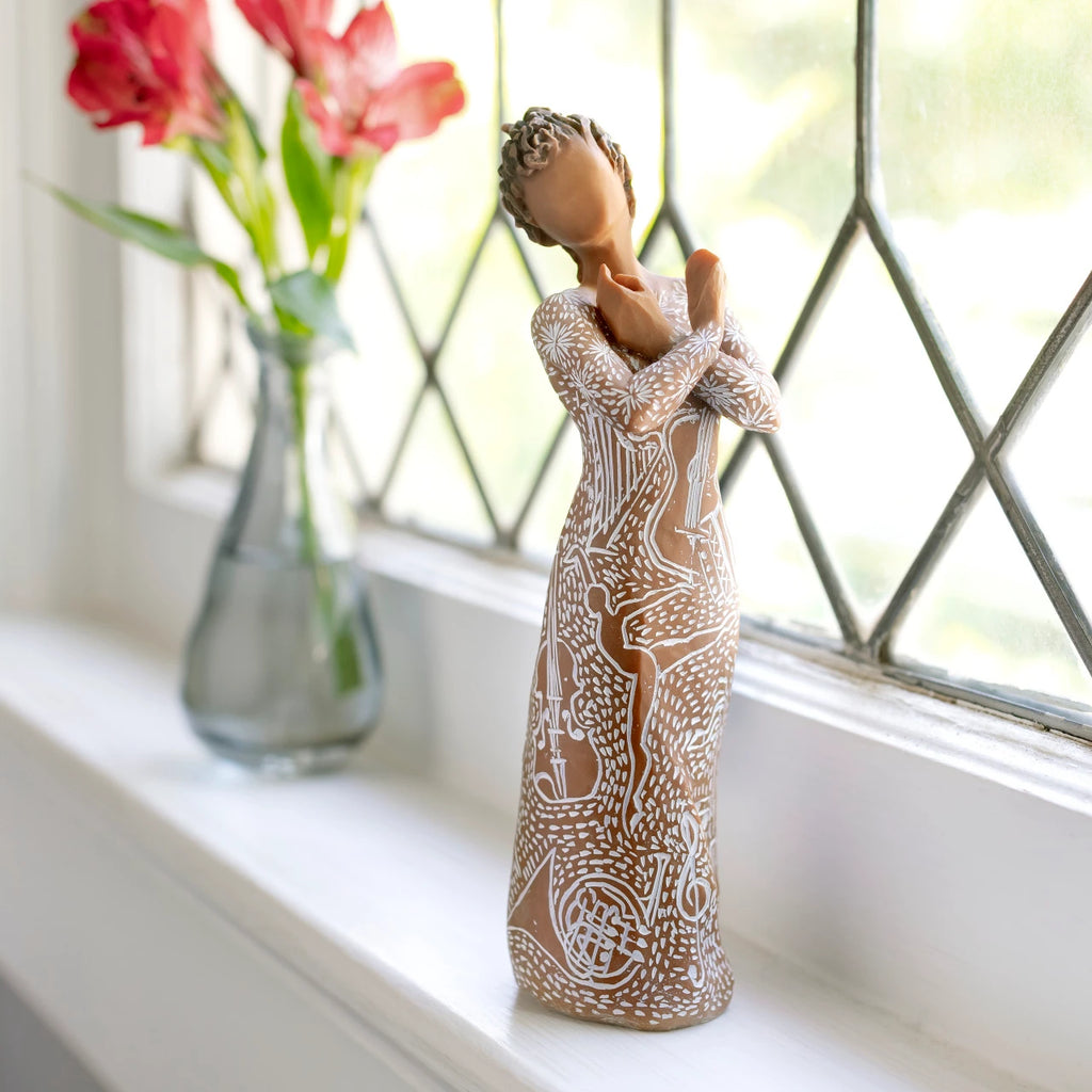 Willow Tree Music Speaks (darker skin), 7”h hand-painted standing resin figure, arms crossed, wearing dress carved with symbols of music and dancing, on bright windowsill